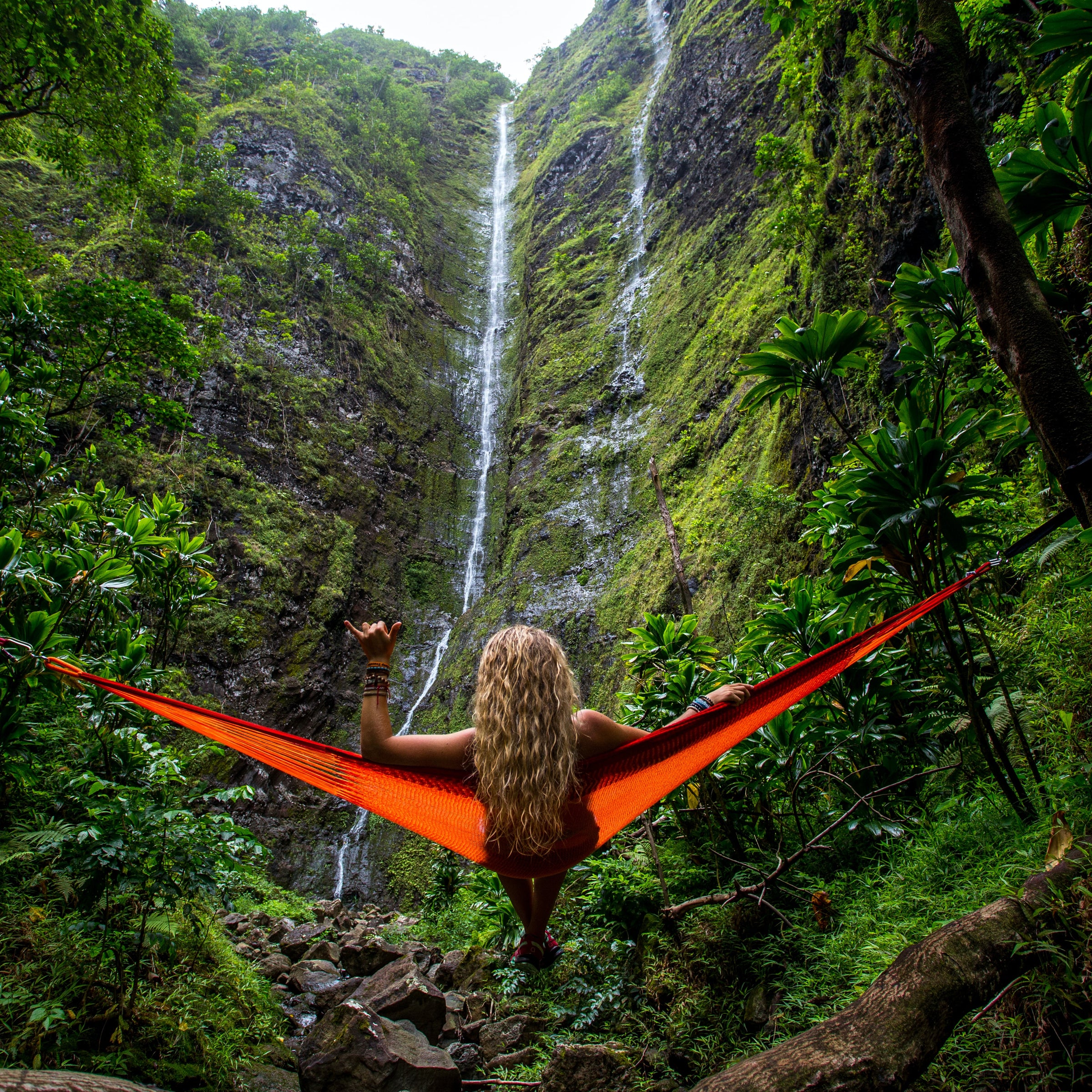 woman giving a shaka sign sitting on a hammock in a very tropical terrain.