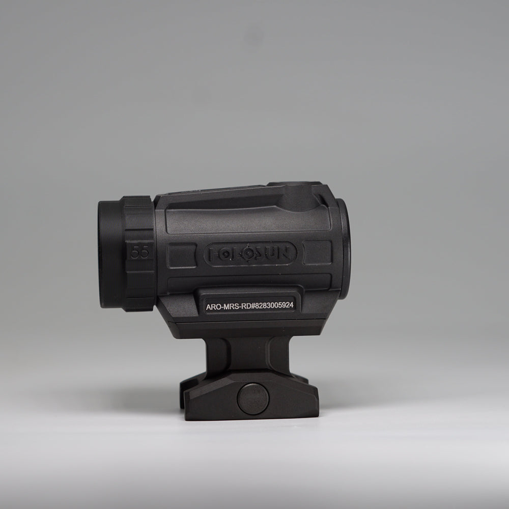 A black tactical-style ARD device with a cylindrical body, perforated front hex pattern on a holosun optic with and mounting base, photographed on a smooth light gray studio background. 