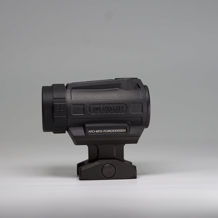 A black tactical-style ARD device with a cylindrical body, perforated front hex pattern on a holosun optic with and mounting base, photographed on a smooth light gray studio background. 