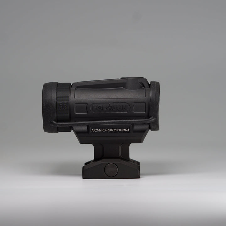 A black tactical-style ARD device on a Holosun Optic with a cylindrical body, perforated front hex pattern, and mounting base, shown using the OM dust cover. Photographed on a smooth light gray studio background. 