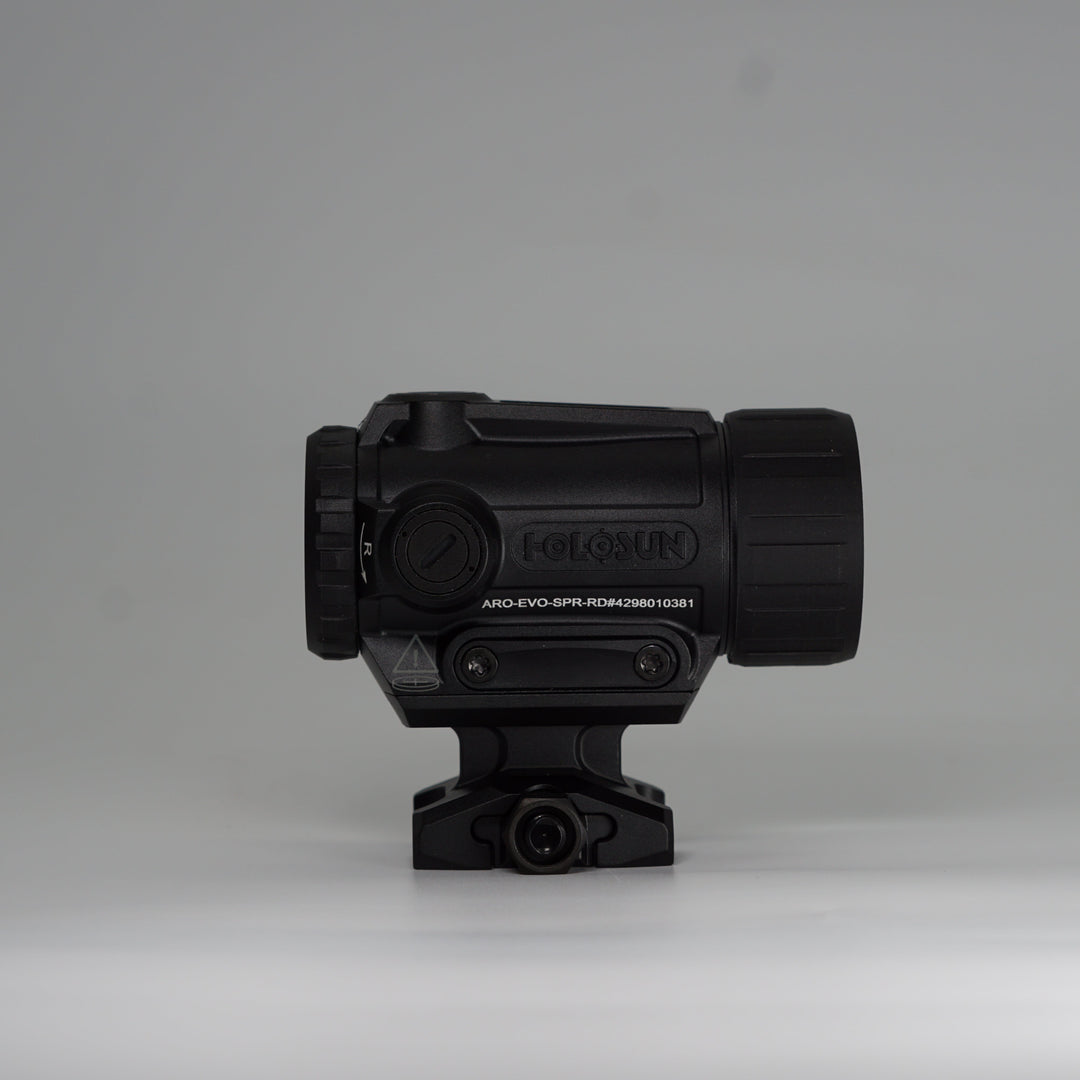 A black tactical-style ARD device with a cylindrical body, perforated front hex pattern, on a holosun ARO red dot optic and mounting base, photographed on a smooth light gray studio background. 