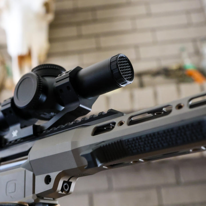 Close-up of a modern rifle with a mounted black scope and a textured black grip, shown against a blurred indoor background with a brick wall and mounted decor.