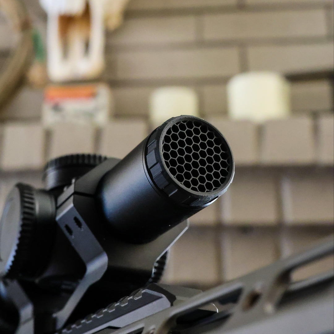 A close-up of a black cylindrical muzzle device or suppressor attached to a firearm, photographed indoors against a blurred brick wall with a mounted animal skull in the background.