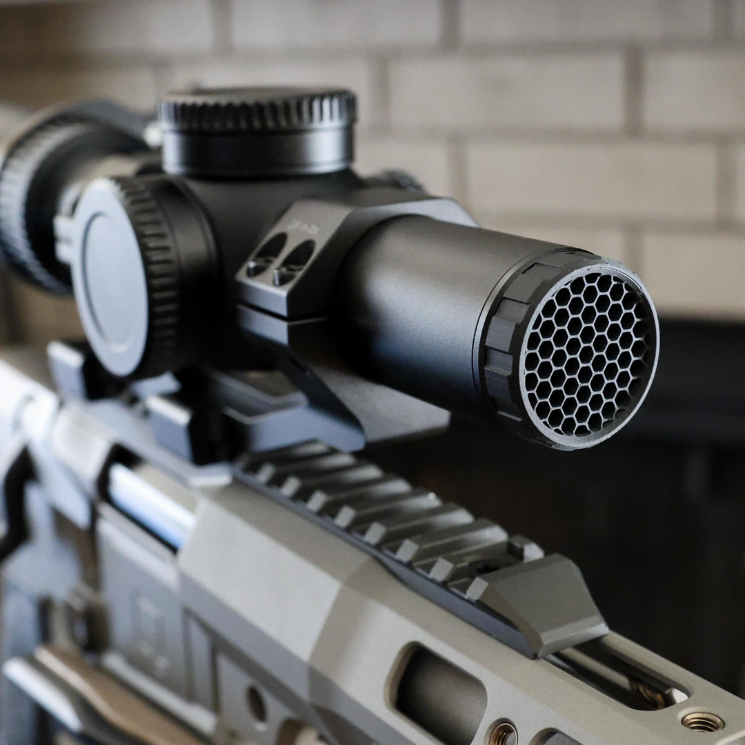 Close-up of a black tactical rifle scope with a honeycomb-patterned ARD device mounted on a Q silver and black rifle, set against a blurred light brick wall background.