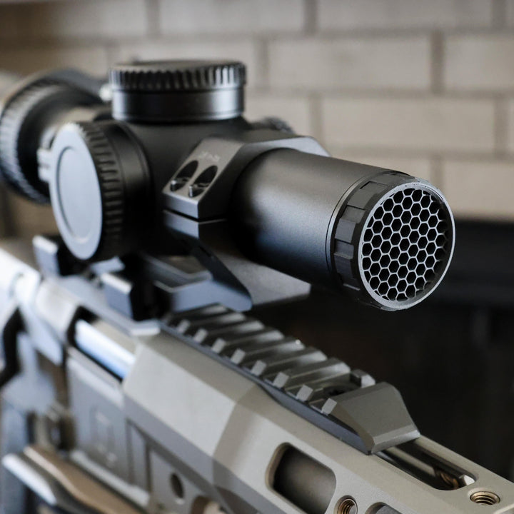 Close-up of a black tactical rifle scope with a honeycomb-patterned ARD device mounted on a Q silver and black rifle, set against a blurred light brick wall background.